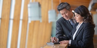 Mature businesswoman and Asian businessman standing on office balcony taking notes.