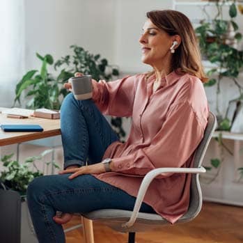 Woman sitting at her desk and listening to meeting on her wireless earphones, while holding a cup of coffee in her hand