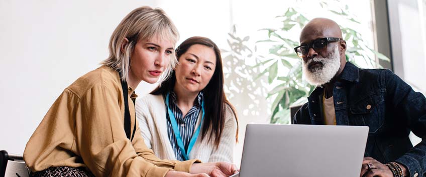 A group of three people - two woman and a man - sit around a laptop, looking at the screen.