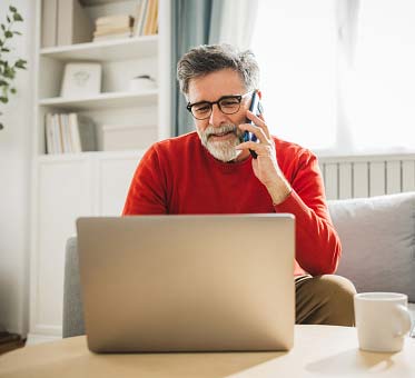 Older man with salt and pepper hair and beard and glasses on his phone in front of a laptop