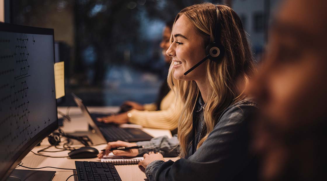 Smiling blond female customer service representative wearing headset using computer at desk in call center